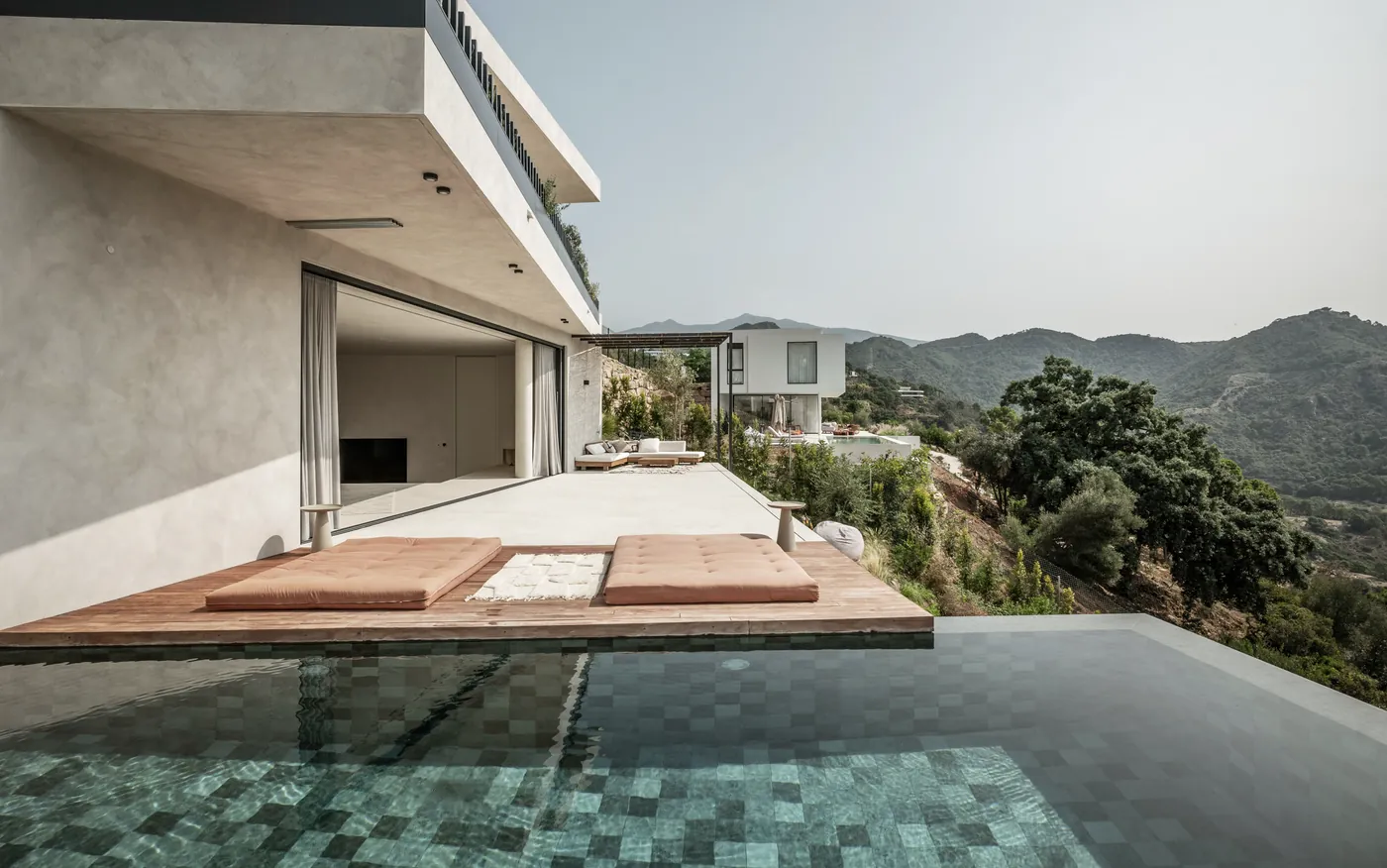 Der Infinity-Pool aus grünem Mosaik unter dem auskragenden Obergeschoss, dahinter die Berge