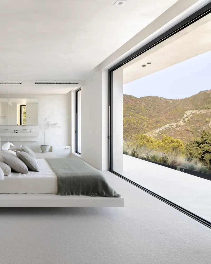 Ein weißes Schlafzimmer mit niedrigem Bett und einer Glaswand zu grünen Bergen